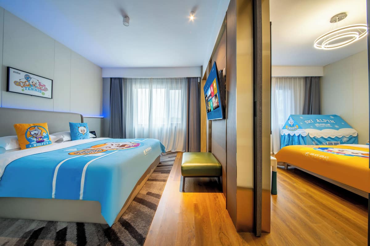 Una habitación de hotel con una cama grande, una cama más pequeña, un banco, un televisor y un ventanal con cortinas.