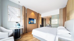A modern hotel room with a large bed, a small round table, a chair, a TV, and a wall-mounted artwork.