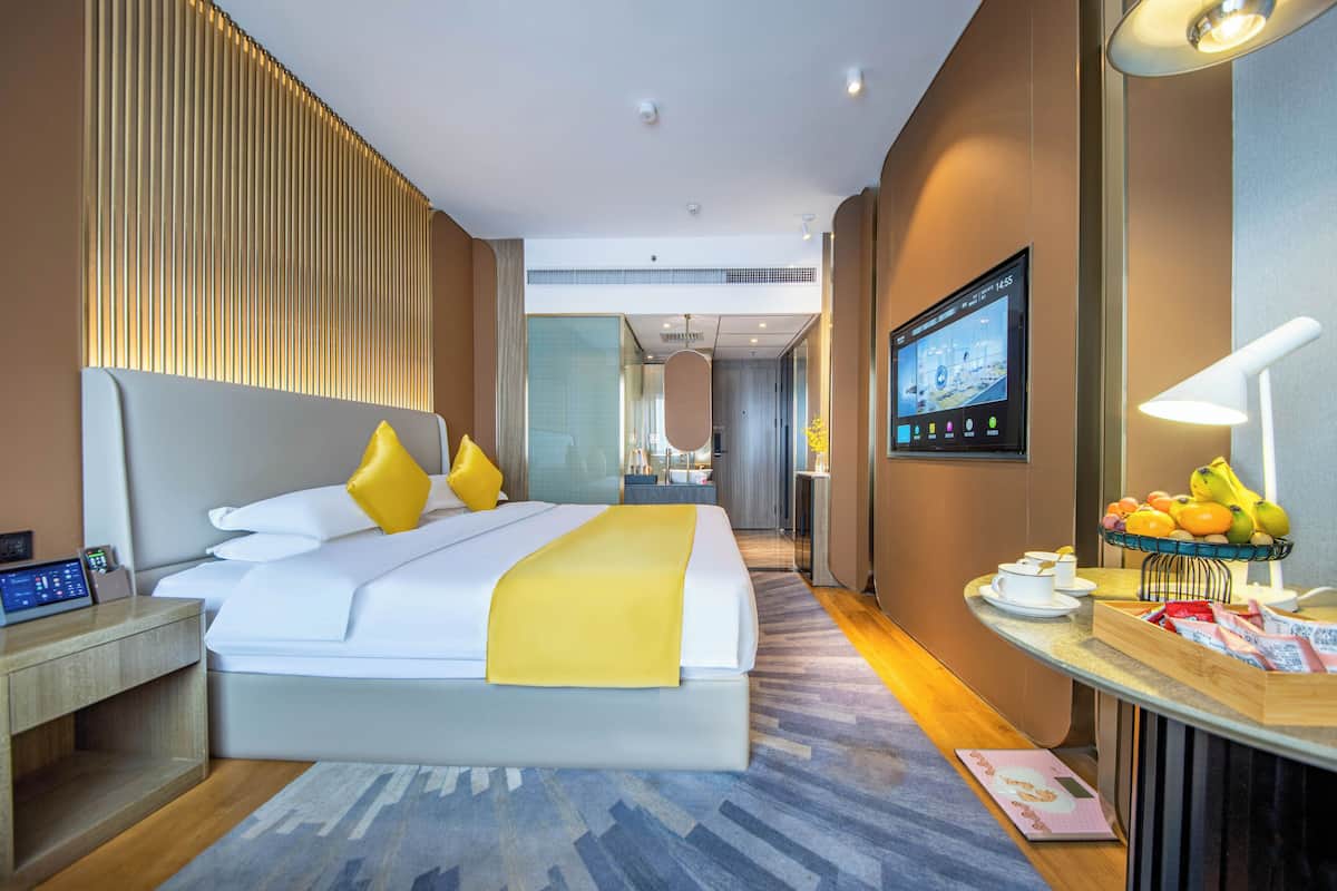 Habitación de hotel con una cama grande, mesita de noche, televisor y una barra de desayuno con fruta.