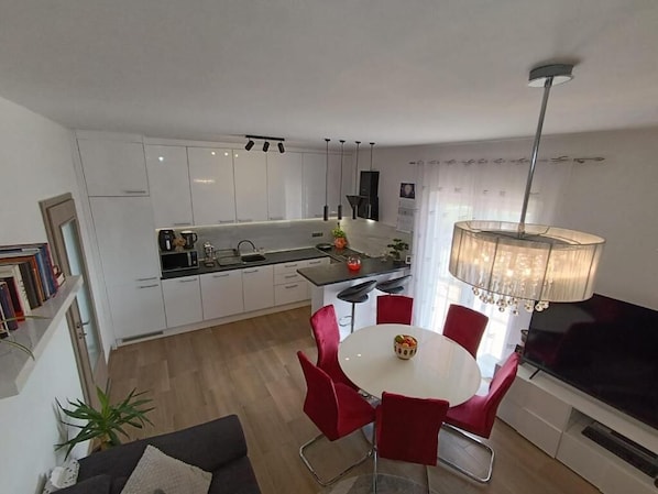 Une cuisine moderne avec des meubles blancs, un coin repas avec des chaises rouges et un grand téléviseur.