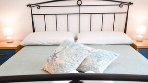 Uma cama com estrutura metálica, almofadas brancas e colcha florida azul. Há duas luminárias de cabeceira e uma mesa pequena com abajur.