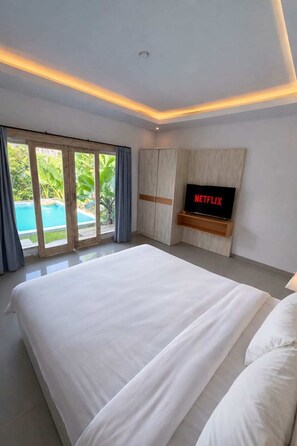 Un dormitorio con una cama grande, un televisor instalado en un panel de madera y un ventanal corredero que da a una piscina.