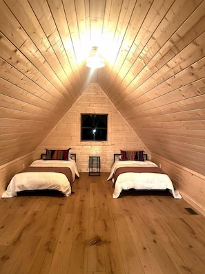 A triangular attic bedroom with wooden walls and floors, two beds, a window, and a light fixture. - Gorgeous A-Frame Cabin Nestled in the Trees near Lost River, West Virginia (Mathias)