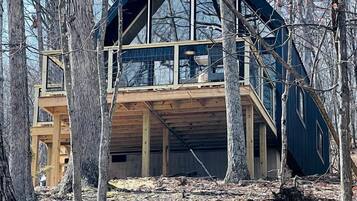 A modern cabin with a large glass facade, wooden deck, and a steeply pitched roof, surrounded by trees.
