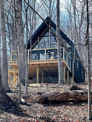 A modern cabin with a large glass facade, wooden deck, and a steeply pitched roof, surrounded by trees. - Gorgeous A-Frame Cabin Nestled in the Trees near Lost River, West Virginia (Mathias)