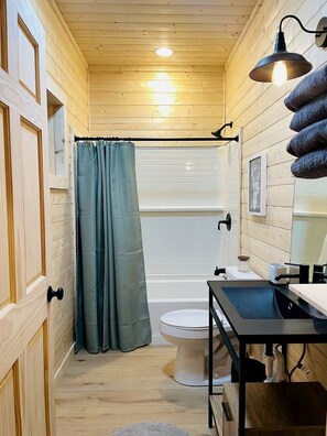 A bathroom with wooden walls, a shower curtain, a toilet, and a sink with a mirror. - Gorgeous A-Frame Cabin Nestled in the Trees near Lost River, West Virginia (Mathias)