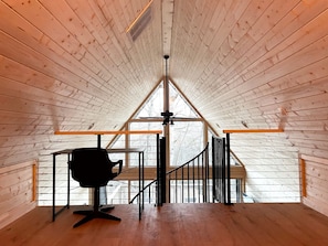Una habitación con techo inclinado, una silla de oficina negra, un escritorio de madera y una escalera que sube a un nivel superior.