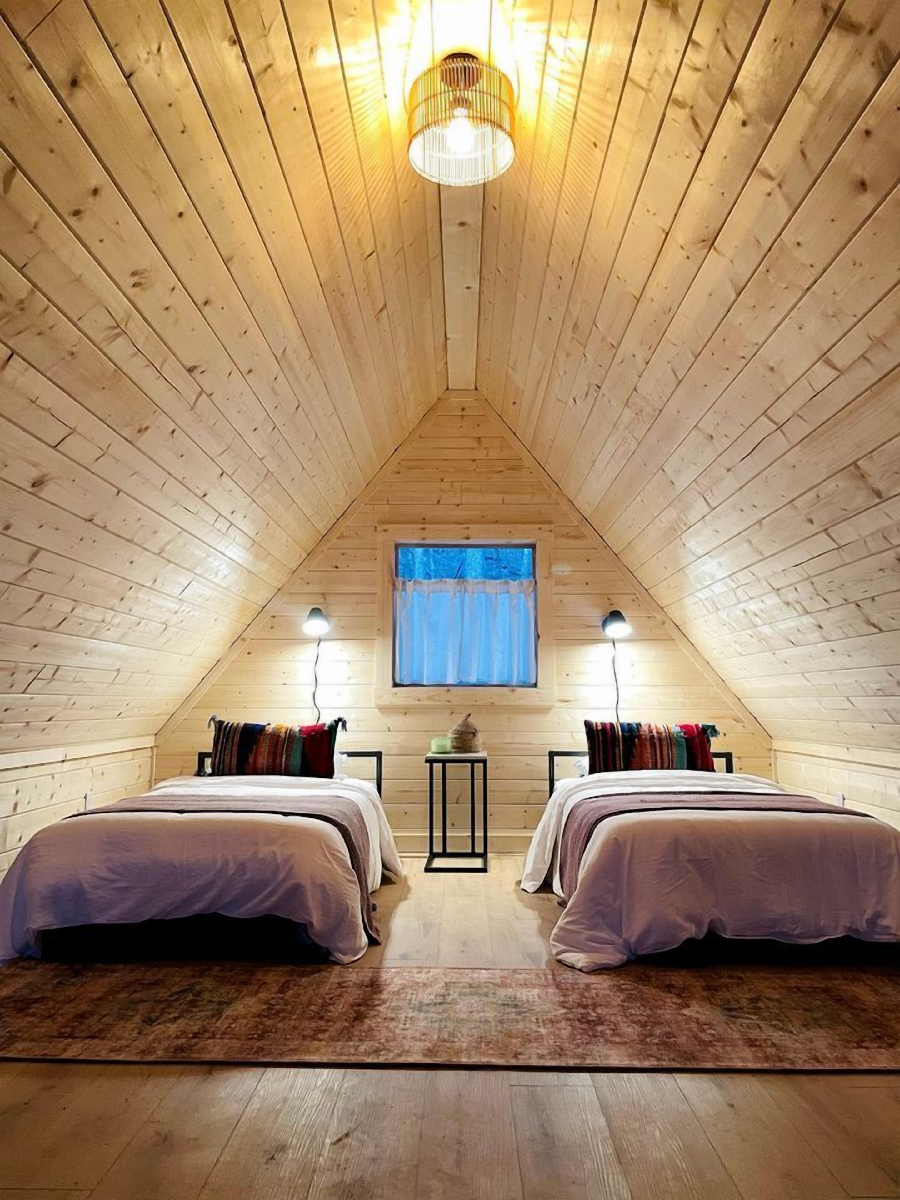 A cozy attic bedroom with two beds, a window with curtains, and a hanging light fixture.