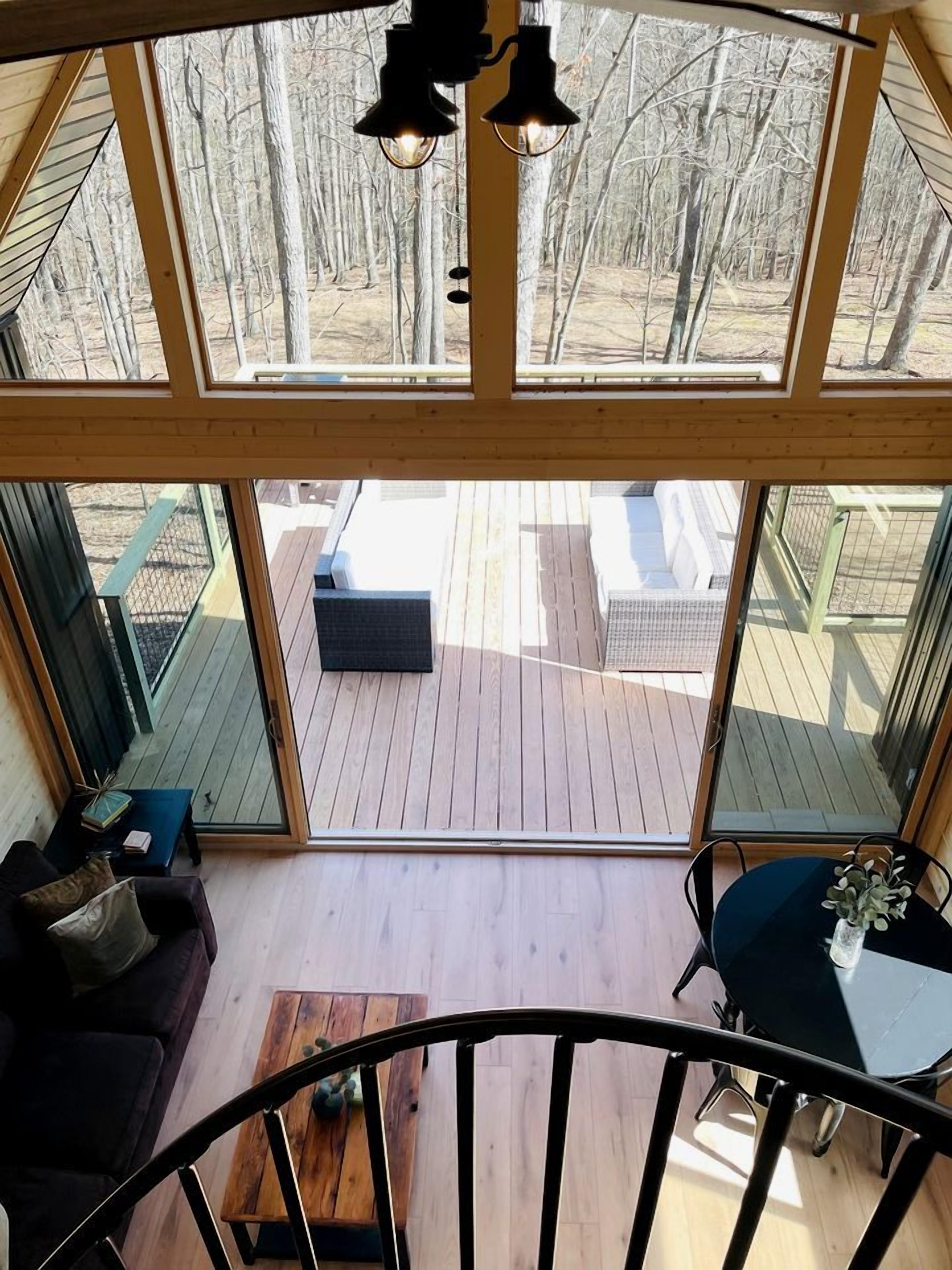 Ein geräumiges Zimmer mit Holzboden, einem dunkelgrauen Sofa, einem kleinen Tisch mit einer Vase und Blick auf eine Holzterrasse und Außenbereich durch große Fenster.
