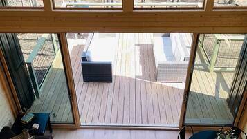 Ein geräumiges Zimmer mit Holzboden, einem dunkelgrauen Sofa, einem kleinen Tisch mit einer Vase und Blick auf eine Holzterrasse und Außenbereich durch große Fenster.