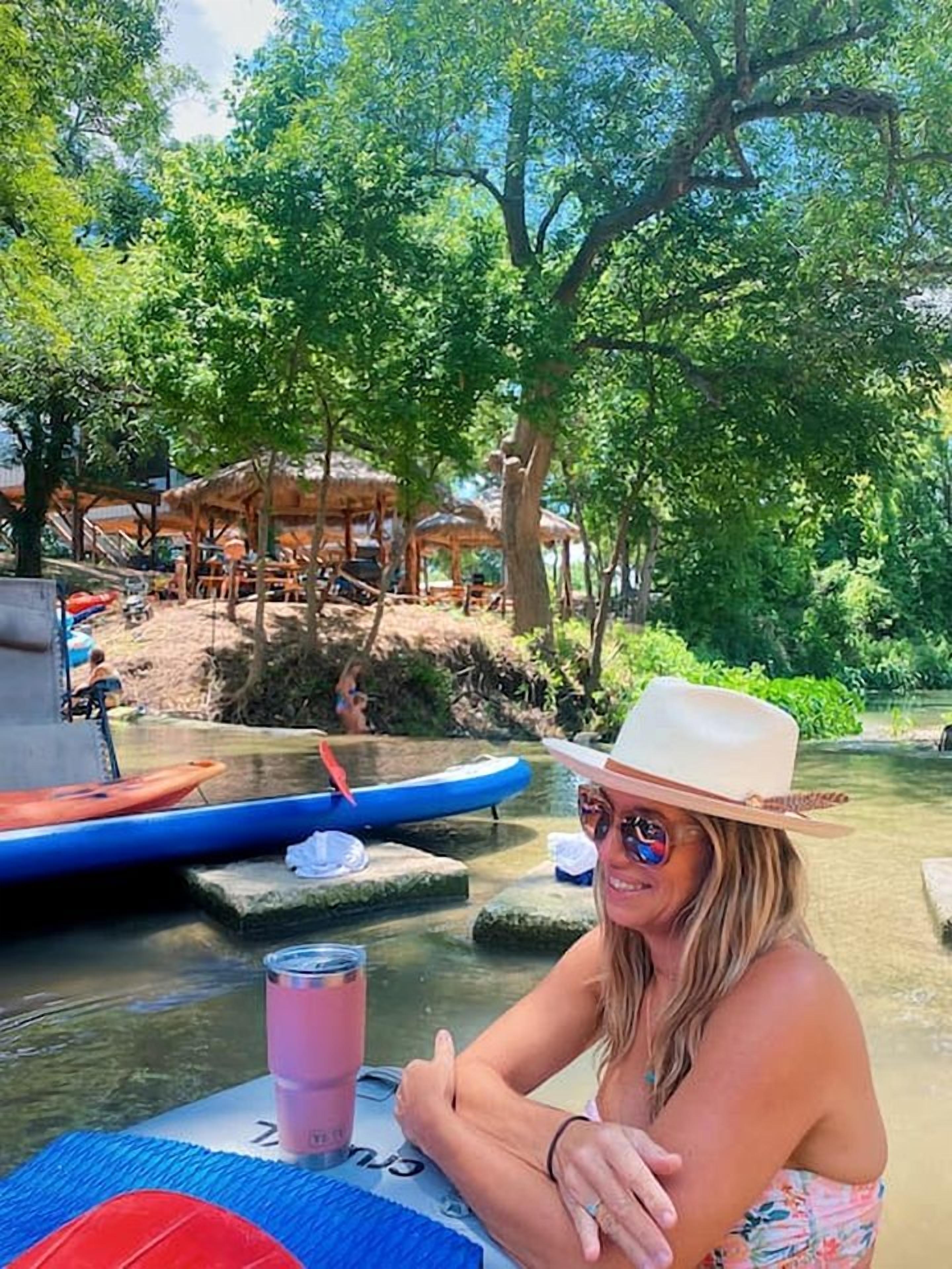 A person seated at a table with a drink, surrounded by a natural setting with trees and a river.