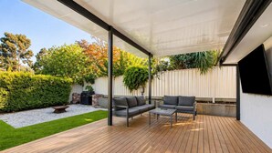 Un patio exterior con terraza de madera, una zona de estar cubierta con sofá y mesa, y una parrilla.