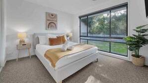 Un dormitorio moderno con un ventanal grande, una cama con colcha color beige, una mesita de noche con lámpara y un cuadro colgado en la pared.