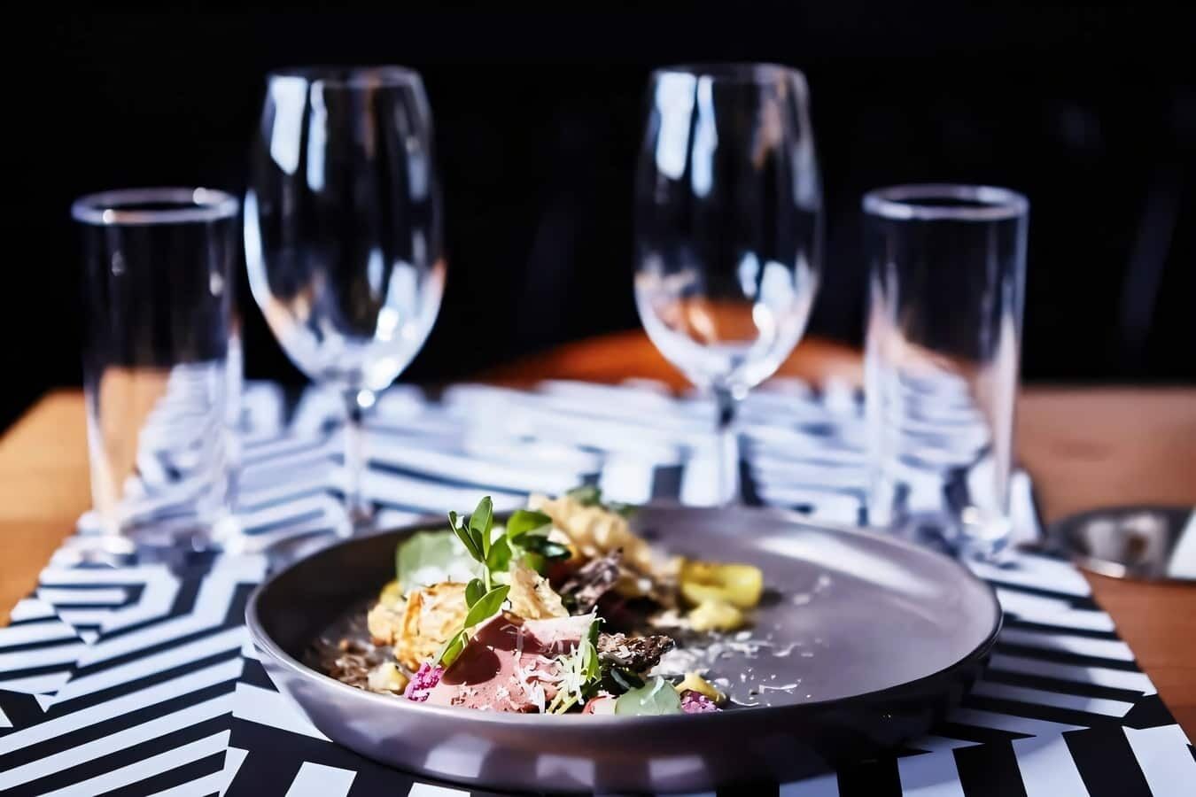 Um prato de comida com toalha de mesa estampada em preto e branco, acompanhado por quatro copos de vinho vazios.