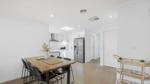 Una cocina moderna con armarios blancos, un refrigerador de acero inoxidable y una zona de comedor con mesa y sillas de madera.