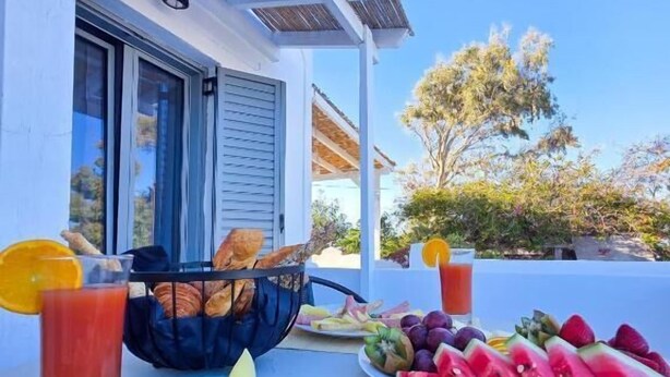 Disposição para o pequeno-almoço com croissants, fruta e sumo numa mesa, em frente a um edifício com vista para árvores e um céu limpo.