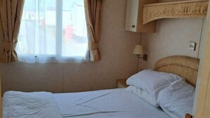 A single bed with a headboard, a window with curtains, a bedside table, and a wall-mounted shelf. - Lyons Sunnysands Holiday Park - Holiday Accommodation 20099 (Talybont)