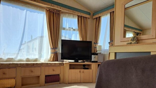 A room with a television on a wooden cabinet, a large window with curtains, and a mirror above the cabinet. - Lyons Sunnysands Holiday Park - Holiday Accommodation 20099 (Talybont)