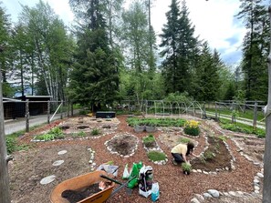 Un jardín con bancales elevados, una carretilla y una persona cuidando plantas.