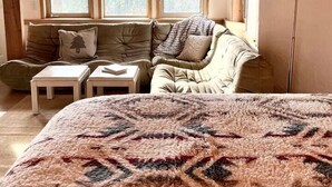 Een gezellige kamer met een bed, een bank, twee kleine tafels en een lamp. Drie ramen bieden uitzicht op het omliggende landschap.
