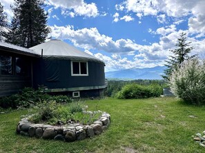A yurt with a stone fire pit, surrounded by a grassy area and trees. - Cozy Mountain Yurt with Panoramic Views near Selkirk Mountains, Sandpoint Idaho (Sandpoint)