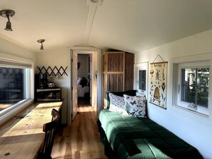 A compact room with a bed, a small kitchenette, a window, and a door leading to another room.