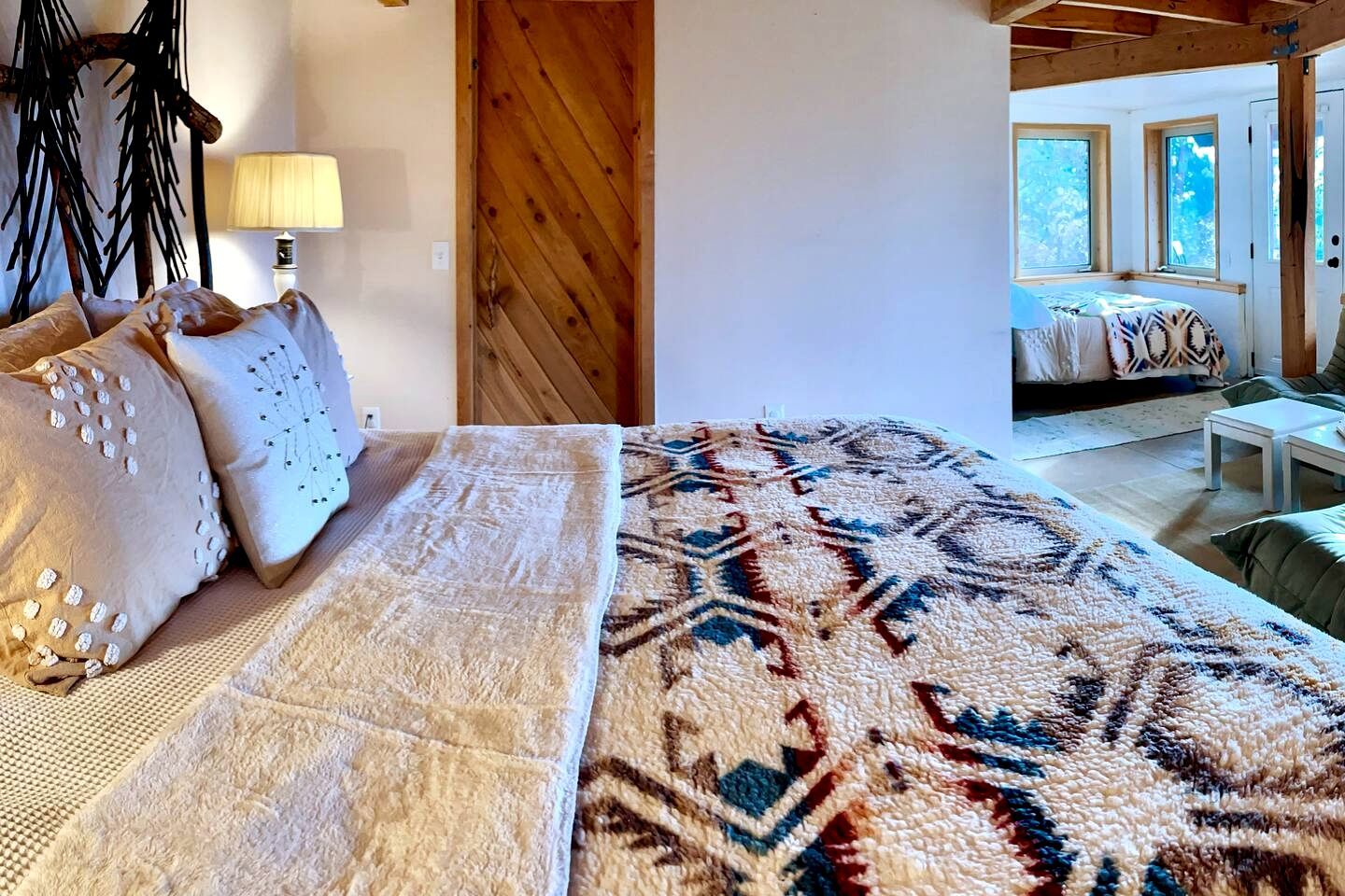 A bedroom with a bed, pillows, a rug, a lamp, and a view of another room with a window.