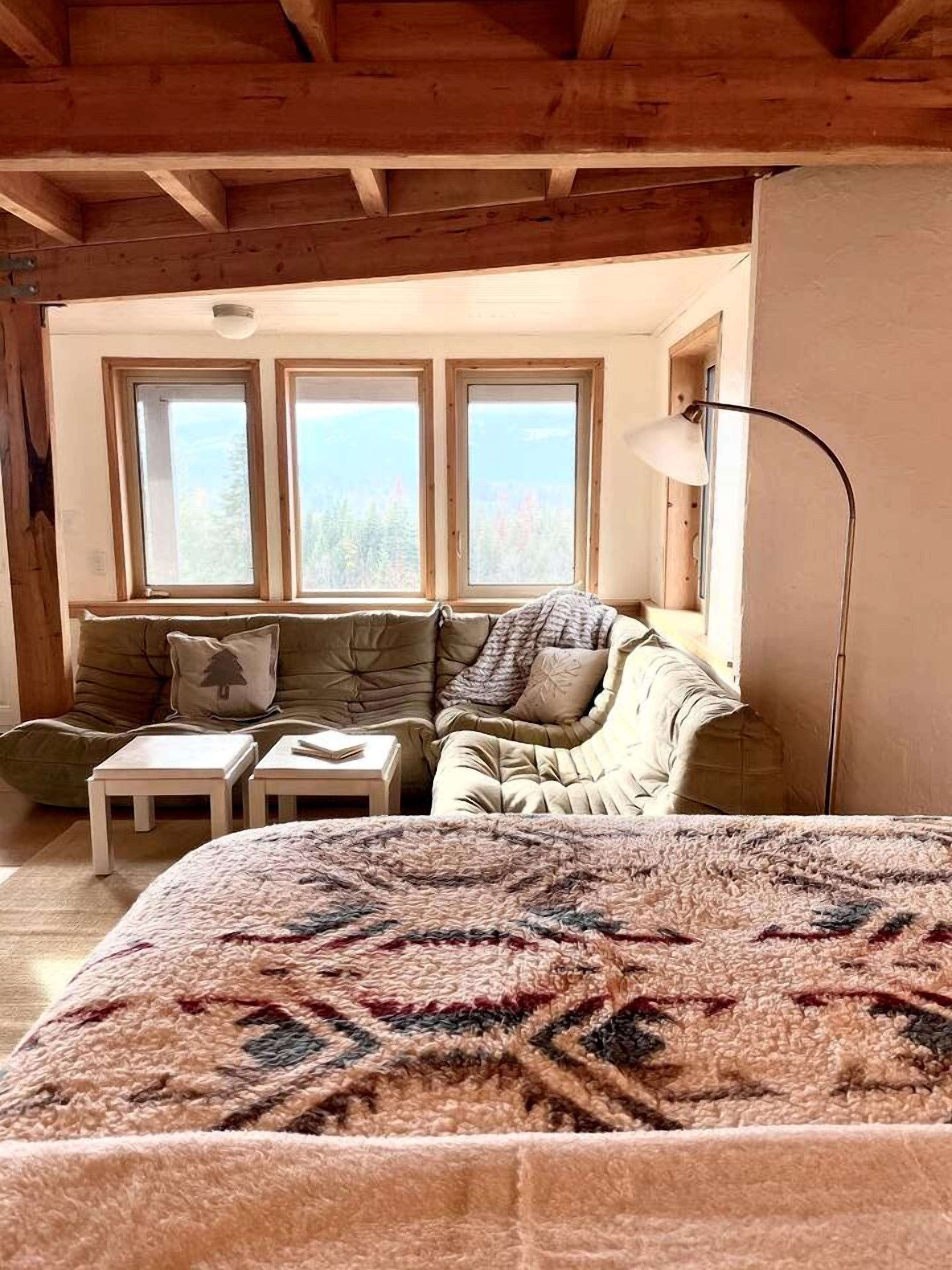A cozy room with a bed, a sofa, two small tables, and a lamp. There are three windows providing a view of the outdoors.