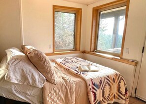 Ein Schlafzimmer mit einem Bett, zwei Fenstern und Blick auf Bäume.