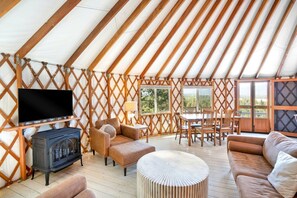 Interior espacioso de una yurta con televisión, asientos, una zona de comedor y ventanas grandes.