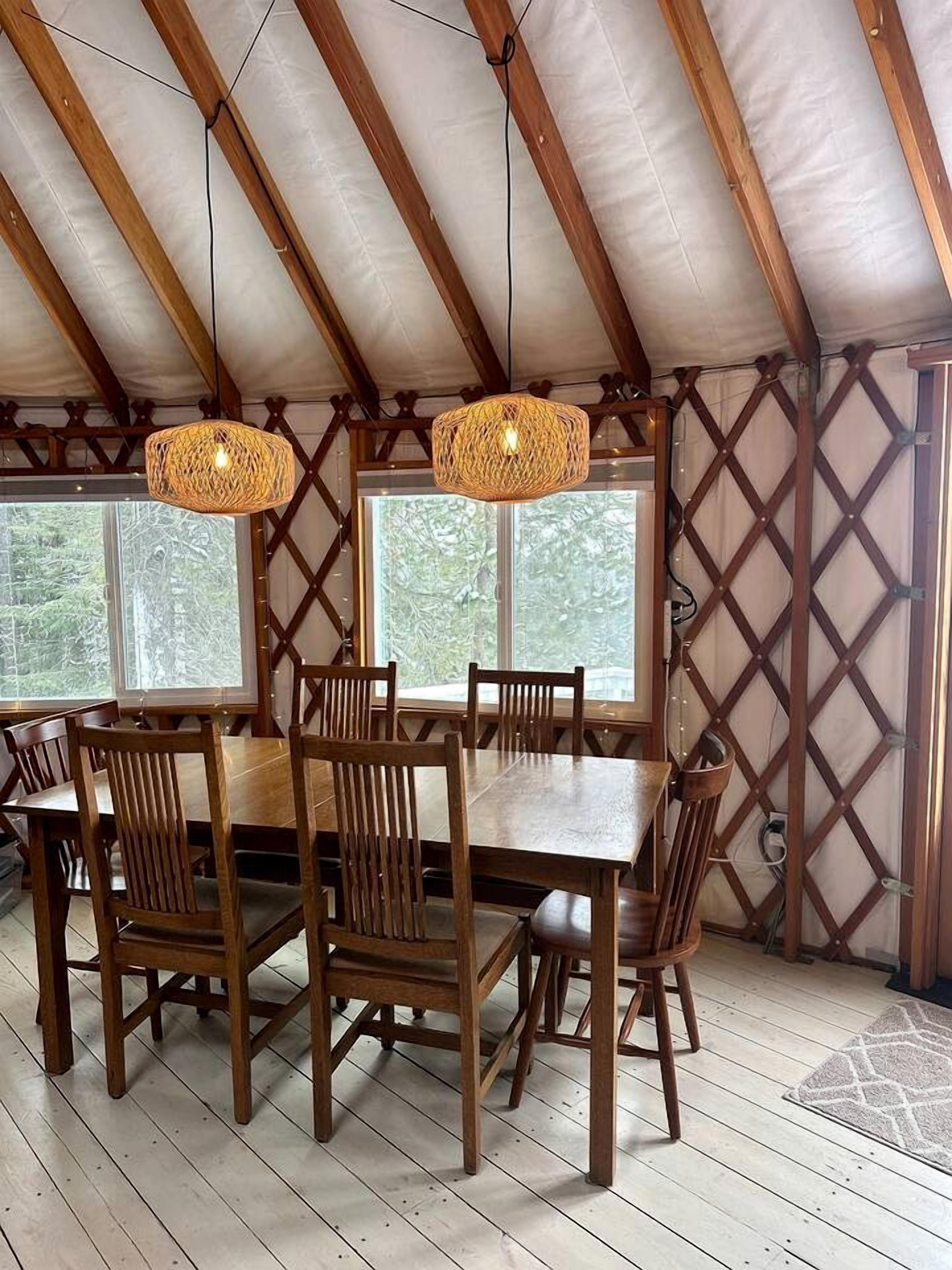 A wooden dining table with chairs, two woven pendant lights, and large windows with a view of trees.