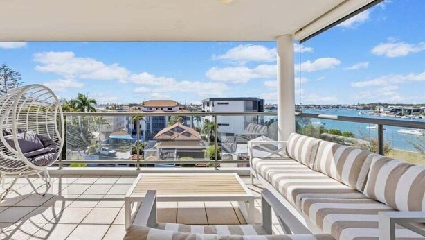A balcony with a sofa, a chair, and a table, overlooking a marina and buildings. - Walk to Beach • Luxury Waterfront Sub-Penthouse (Sunshine Coast)