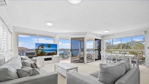 A modern living room with a large window, a sofa, a coffee table, and a dining area with chairs. - Walk to Beach • Luxury Waterfront Sub-Penthouse (Sunshine Coast)