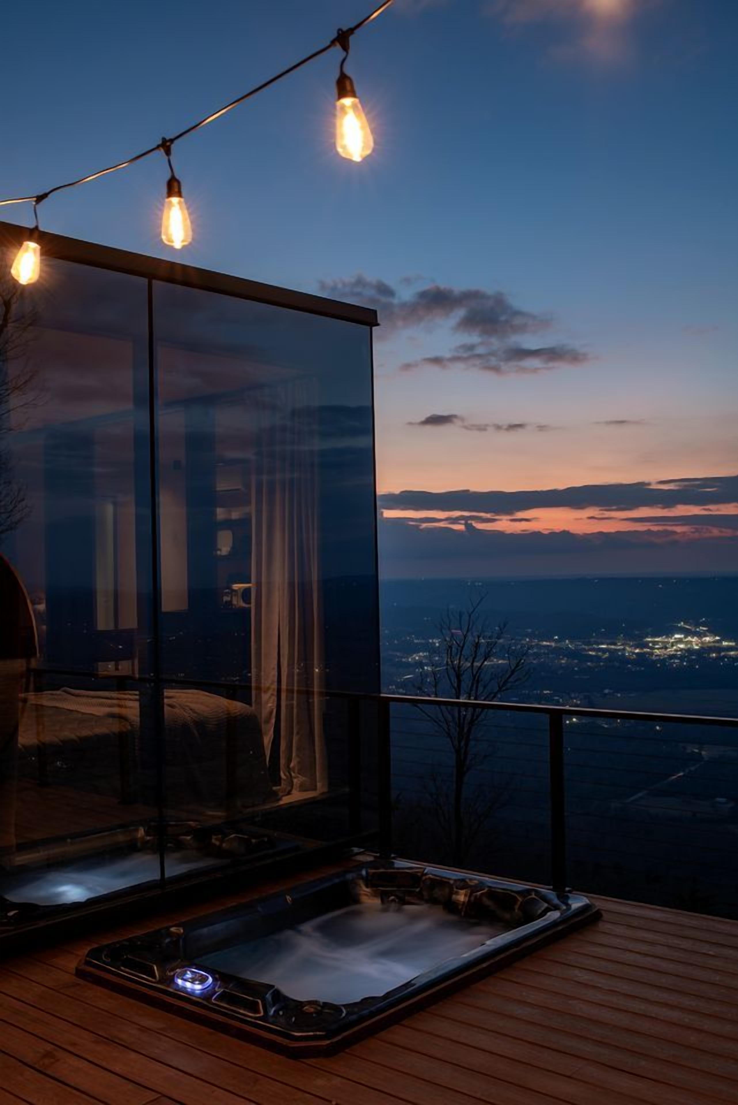 A modern room with a glass wall, a hot tub, and a city view at dusk.