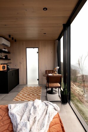 Ein modernes Interieur mit großem Fenster, einer Küchenzeile, einer Sitzecke mit Tisch und Stühlen sowie einem Bett mit Decke.