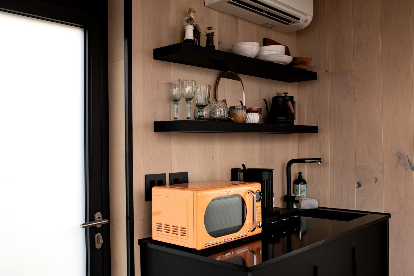 A compact kitchen with a microwave, shelves with dishes and glasses, and a coffee maker.