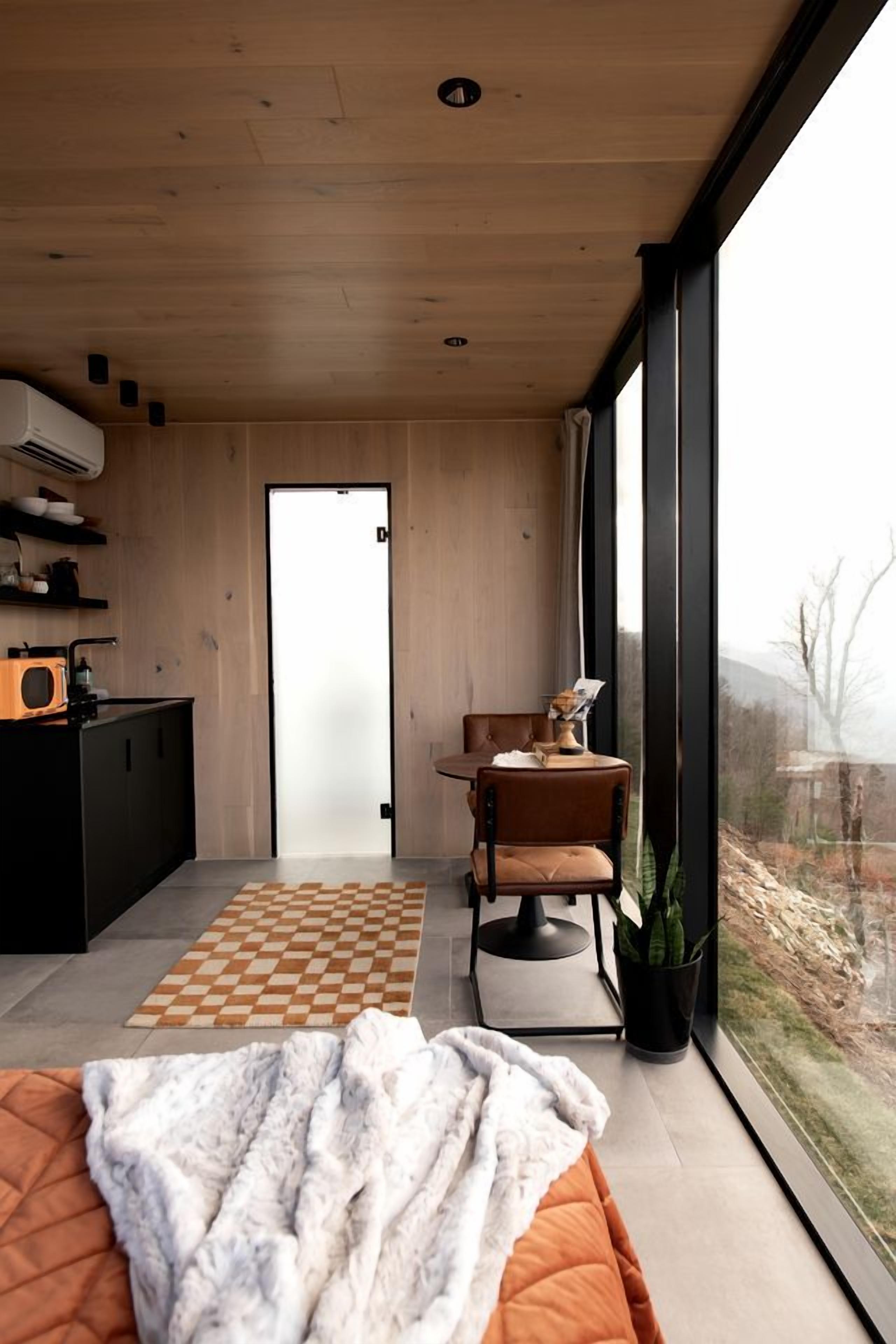 A modern interior with a large window, a kitchen area, a seating area with a table and chairs, and a bed with a blanket.