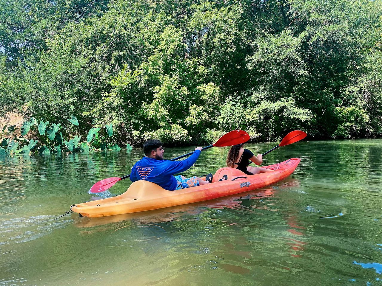 Zwei Personen paddeln in einem ruhigen Fluss, umgeben von dichtem Grün.