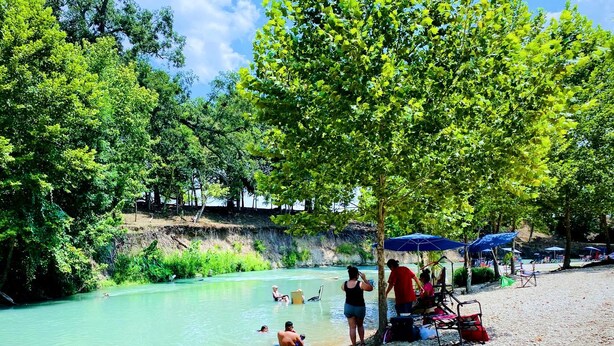 Una sponda di fiume con persone che nuotano e si rilassano, circondata da alberi e ombrelloni.
