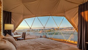 A tented room with a bed, a table, and a chair, and a view of a landscape with mountains and a river.
