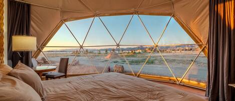 A tented room with a bed, a table, and a chair, and a view of a landscape with mountains and a river.