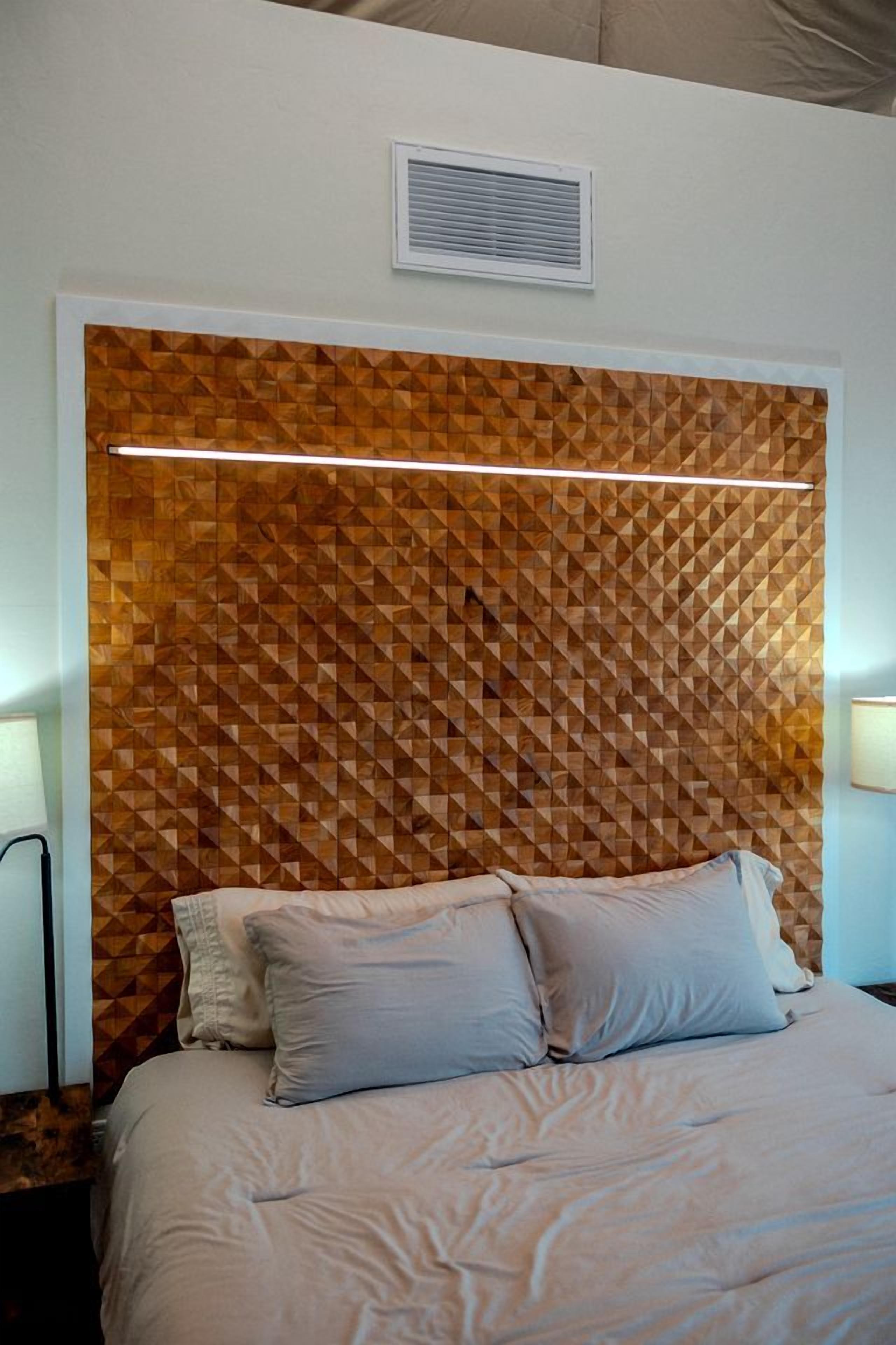 Um quarto com cabeceira estampada em padrão geométrico, uma cama com roupa de cama em tons claros e uma mesa de cabeceira com luminária.