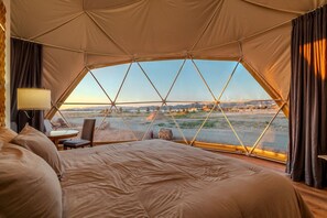 A tented room with a bed, a table, and a chair, and a view of a landscape with mountains and a river.