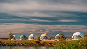 Une rangée de structures en forme de dômes géodésiques, situées au bord d’un plan d’eau, sous un ciel nuageux.