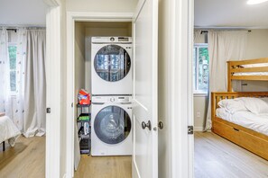 Quarto compacto com máquina de lavar, cama e prateleira pequena com produtos de limpeza.
