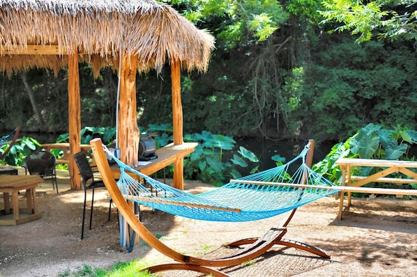A hammock, wooden chairs, a table, and a thatched-roof structure in a garden setting.