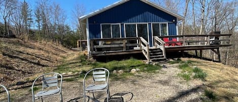 A blue cabin with a deck, surrounded by trees and a gravel area with metal chairs.