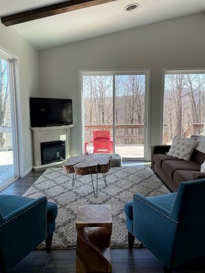 A living room with a fireplace, a television, a large window, and a view of trees.