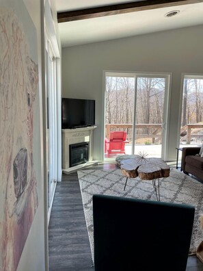 A modern living room with a fireplace, a flat-screen TV, a large window, and a view of the outdoors. - Relaxing Converted Barn Surrounded by Nature near Lost River, West Virginia (Mathias)