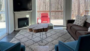 A living room with a fireplace, a television, a large window, and a view of trees.